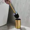Brosse WC or avec poignée dorée et brosse noire, dans son support doré près d'un WC.