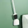 Brosse WC blanche avec poignée longue et poils noirs, fixée sur un porte-brosse mural près d'un WC.